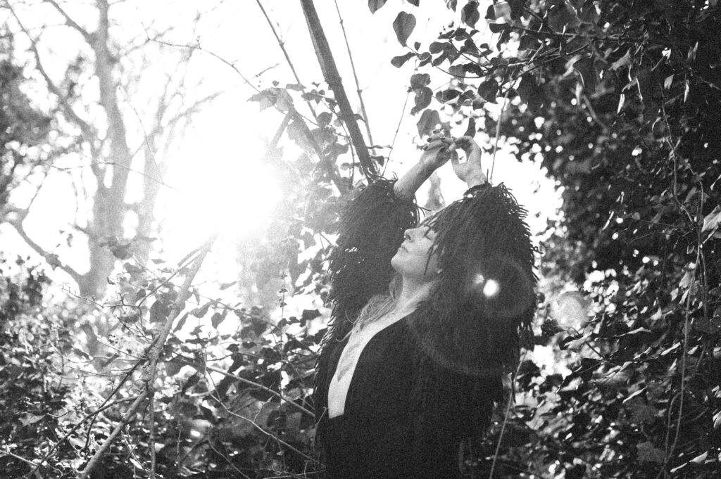 Black and white photo of Fiona McCoss with her arms in the air and eyes closed, basking in the sunlight amongst the trees. Blog post: Are you ready to step into Feminine Leadership?
